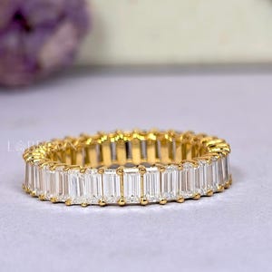 May include: A gold eternity ring with rectangular-cut, clear gemstones. The ring is set with multiple stones, creating a continuous band of sparkle. The ring is displayed on a light-colored surface, with a blurred floral background.