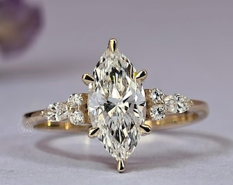 Könnte beinhalten: Ein goldener Verlobungsring mit einem Diamanten im Marquise-Schliff als Mittelpunkt. Der Ring hat kleinere Diamanten auf beiden Seiten des Hauptsteins und entlang des Bandes. Der Ring ist vor einem weichen, neutralen Hintergrund platziert, der das Funkeln der Diamanten hervorhebt.