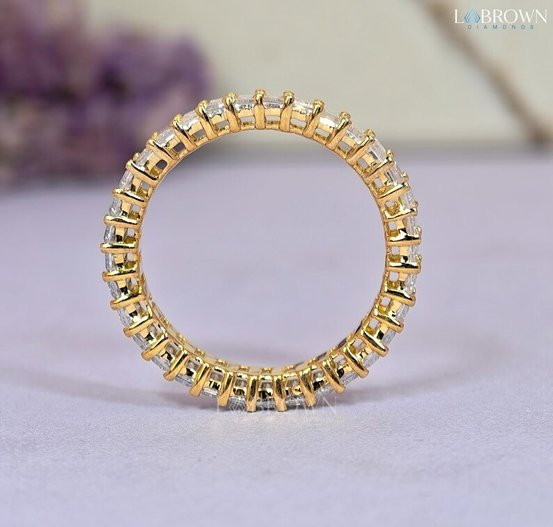 May include: A gold eternity ring with a continuous row of rectangular-cut, clear gemstones. The ring is set in a yellow gold band, creating a sparkling and elegant design. The ring is displayed on a light purple surface.