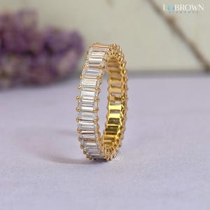 May include: A gold band ring with a continuous row of rectangular, clear gemstones. The ring is set on a light purple surface, with a blurred background of purple flowers and a white surface.