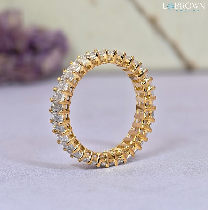 May include: A gold eternity ring with a continuous row of rectangular-cut diamonds. The ring's band is a warm gold color, and the diamonds are clear and sparkling. The ring is a classic piece of jewelry.