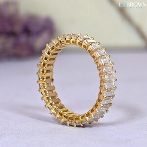 May include: A gold eternity ring featuring a continuous row of rectangular-cut, clear gemstones. The ring's band is a warm gold color, and the gemstones are set closely together, creating a sparkling effect. The ring is a classic piece of jewelry.