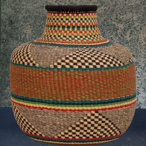 May include: A handwoven basket with a wide base and a narrow neck. The basket has a geometric pattern in black, white, red, green, and brown. The rim of the basket is black.