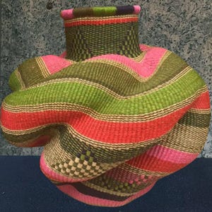 Puede incluir: Una cesta tejida con un diseño anudado único. La cesta presenta rayas horizontales en tonos de rosa, verde, rojo y marrón. La parte superior tiene un patrón de cuadros. La cesta es un objeto decorativo.