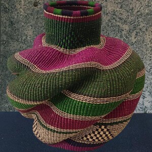 May include: A decorative woven basket with a distinctive, swirling form. The basket is made with bands of green, magenta, and natural-toned fibres. The top rim is a darker green and purple. The base has a checkered pattern.