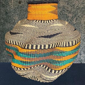 Puede incluir: Una cesta tejida a mano con un cuerpo bulboso y un cuello estrecho. La cesta presenta bandas horizontales de color naranja, turquesa y negro, con un fondo crema. El cuello es naranja con diseños de chevrón negro. La cesta está hecha a mano.