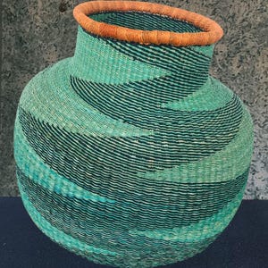 Puede incluir: Una cesta tejida con base redondeada y cuello más estrecho. La cesta presenta un patrón de rayas turquesa y negro, con un borde naranja. La textura de la cesta es visible, mostrando las fibras naturales utilizadas en su construcción.