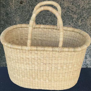 Puede incluir: Una cesta tejida de forma ovalada con dos asas arqueadas. La cesta está hecha de material tejido de color beige claro. Las asas también están hechas del mismo material y están unidas al borde superior de la cesta.