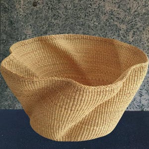 Puede incluir: Una cesta grande y tejida con un diseño curvo único. La cesta está hecha de fibras de color natural, con una superficie texturizada y una parte superior ancha y abierta. La forma de la cesta es asimétrica, con una forma fluida y orgánica. La cesta es adecuada para almacenamiento o exhibición.