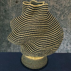 May include: A decorative woven basket with a distinctive, organic form. The basket features black and natural beige horizontal stripes. It has a round base and a flared, ruffled top. The basket is crafted from natural fibres and would suit any home.