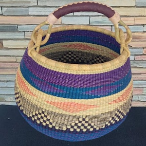 Puede incluir: Cesta redonda tejida a mano con asa. La cesta presenta rayas horizontales en morado, azul, melocotón y crema. El asa está envuelta en material morado oscuro. La base tiene un patrón de cuadros. Un artículo de almacenamiento decorativo.