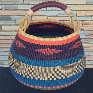 Puede incluir: Cesta tejida a mano, de forma redonda, con asa. La cesta presenta un diseño vibrante con bandas azules, naranjas, moradas y beige. El asa está envuelta en cuero marrón oscuro. Adecuada para almacenamiento o decoración. Diámetro aproximado de 30 cm.