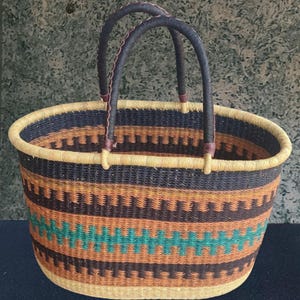 Puede incluir: Cesta ovalada tejida con un patrón geométrico repetido en negro, naranja y turquesa. La cesta tiene dos asas marrones oscuras con detalles de cuero y un borde de color beige claro. La cesta mide aproximadamente 35 cm de ancho y 25 cm de alto.