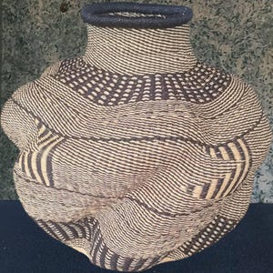 May include: A handwoven basket with a distinctive form, wide at the base and tapering to a narrow neck. It's crafted from natural fibres, displaying a pattern of alternating dark and light stripes. The rim is a solid dark colour.