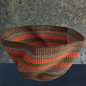 Puede incluir: Una cesta tejida con una abertura amplia. Presenta rayas horizontales en naranja, verde y blanco, con un borde y base marrones. Hecha de fibras naturales, la cesta tiene una forma ligeramente irregular, resaltando su calidad artesanal.