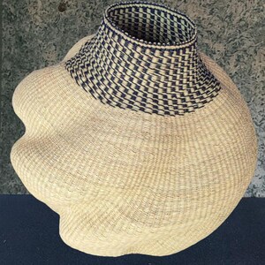 May include: A woven basket with a distinctive form. The main body is a light beige, with a navy blue and white geometric pattern on the neck. The basket has a broad, rounded base and a flared neck, likely for storage.