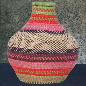 Könnte beinhalten: Ein gewebter Korb mit einem bauchigen Körper und einem schmalen Hals. Der Korb hat horizontale Farbstreifen, darunter Rot, Rosa, Grün und Braun. Der obere Rand ist grün.