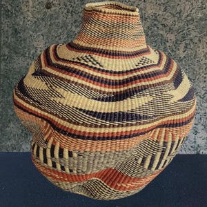 May include: A handwoven basket with a rounded base and a slender neck. The basket displays a geometric design with alternating stripes of beige, red, brown, and navy blue. The woven texture is clearly visible.