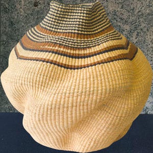 Puede incluir: Cesta tejida con un cuerpo bulboso y un cuello estrecho. La cesta presenta bandas horizontales de fibras naturales, marrones y azul oscuro. El cuerpo tiene una forma texturizada y redondeada, y la cesta está hecha de material tejido de color natural.