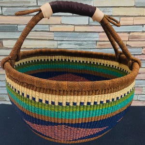 Puede incluir: Una cesta tejida a mano con un asa de cuero marrón y un diseño a rayas colorido. La cesta presenta una mezcla de tonos azules, verdes, rosas y beige, creando una apariencia vibrante y texturizada. Ideal para almacenamiento o decoración.