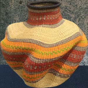 May include: A woven basket with a wide, rounded body and a narrow neck. The basket features horizontal bands of colour, including beige, orange, red, and green, with a dark brown rim and accents. The basket is made of natural fibres.