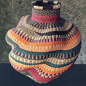 Puede incluir: Una cesta tejida a mano con una forma única y bulbosa y un cuello estrecho. La cesta presenta una vibrante gama de colores, incluyendo naranja, turquesa, rosa y negro, dispuestos en bandas horizontales. La textura del material tejido es claramente visible.