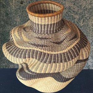 May include: A hand-woven basket with an unusual, organic form. It has alternating bands of dark brown and natural beige fibres, creating a striped pattern. The rim is a darker brown, and the base is a lighter beige.