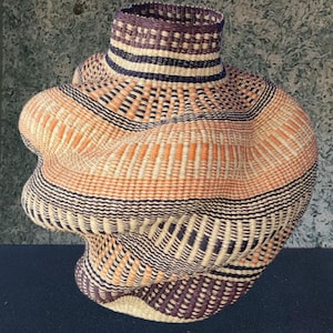 May include: A handwoven basket with a distinctive, rounded form and a slender neck. The basket showcases an intricate pattern of woven fibres in beige, orange, black, and purple hues. The texture and design highlight craftsmanship and artistic skill.