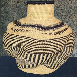 May include: A hand-woven basket with a distinctive form, wide at the base and tapering to a narrow neck. It's crafted from natural-toned fibres, adorned with horizontal bands of dark blue or black. The neck's rim is also dark blue.