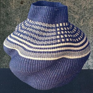 May include: A handwoven basket with an unusual, organic form. The basket is mainly navy blue, with a pattern of cream-coloured horizontal and curved stripes. The basket has a broad, rounded base and a narrower, cylindrical neck.