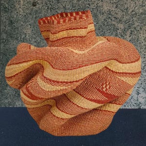 Puede incluir: Cesta tejida de forma única, con bandas alternas de rojo y beige natural. La cesta tiene una superficie texturizada y una parte superior abierta y plegada. El diseño incluye un sutil patrón tejido en el lateral. Esta cesta decorativa es adecuada para almacenamiento o exhibición.