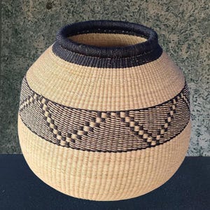 Puede incluir: Una cesta tejida con forma redondeada, con un cuerpo beige natural y un borde y banda decorativa azul oscuro. La banda tiene un patrón geométrico. La cesta es probablemente hecha a mano y adecuada para almacenamiento o decoración.