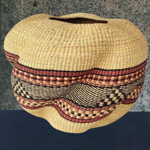 May include: A hand-woven basket with an unusual, organic form. The basket is made of natural-coloured fibres with a decorative band of red, black, and cream-coloured woven patterns. The basket has a wide opening at the top.
