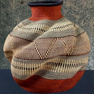 Puede incluir: Una cesta tejida con un cuerpo bulboso y un cuello estrecho. La cesta presenta bandas horizontales de varios colores, incluyendo rojo, marrón, beige y gris. El borde es azul oscuro. La cesta tiene una superficie texturizada y una apariencia artesanal única.