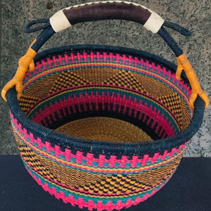 Puede incluir: Una cesta redonda tejida con un borde y soportes de asa azul oscuro. La cesta presenta un vibrante patrón de rayas horizontales rosas, amarillas, negras y turquesas. El asa es de cuero marrón con detalles blancos y soportes tejidos amarillos.