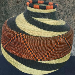 May include: A hand-woven basket with a wide, rounded body and a narrow neck. The basket features a repeating pattern of black, orange, and natural-coloured woven bands. The rim of the basket is striped with orange and black.