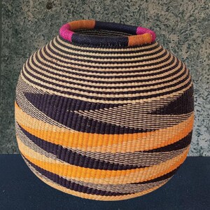 Könnte beinhalten: Ein gewebter Korb mit runder Form, mit horizontalen Streifen in Beige und Schwarz, mit diagonalen Bändern in Orange und Marineblau. Der Rand ist mit einem Band aus Pink, Orange und Marineblau akzentuiert. Dieser dekorative Korb eignet sich zur Aufbewahrung oder Präsentation.