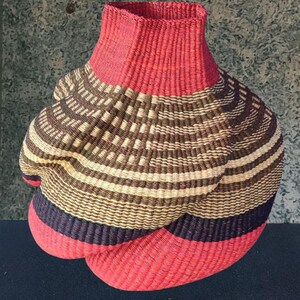 May include: A woven basket with a red rim and base, and a patterned body. The body features alternating bands of brown, tan, and black, creating a textured design. The basket has a unique, bulbous shape.