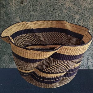 Puede incluir: Cesta tejida con una forma única. La cesta presenta bandas alternas de color beige natural y azul oscuro. El tejido incluye un diseño geométrico. La cesta está hecha de fibras naturales y tiene una construcción robusta.