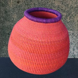 Puede incluir: Una cesta tejida a mano con un cuerpo redondeado y una abertura amplia. La cesta es principalmente de color coral vibrante, con un borde morado oscuro alrededor de la abertura. La textura del material tejido es claramente visible.