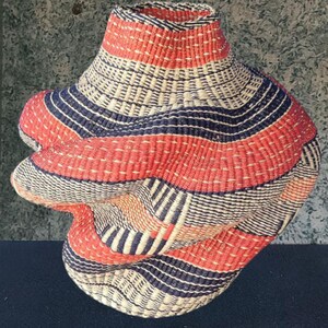 May include: A handwoven basket with a distinctive, sculptural form. It displays horizontal bands of red, navy blue, and natural beige fibres. The design includes a flared top and a bulbous, asymmetrical body, showcasing intricate weaving patterns.