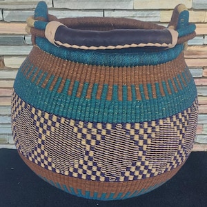 Puede incluir: Una cesta tejida a mano de forma redonda con una abertura ancha. Presenta patrones tejidos en marrón, turquesa y morado. Tiene dos asas y un borde envuelto en cuero azul oscuro. La cesta, de unos 30 cm de diámetro, es adecuada para almacenamiento o decoración.