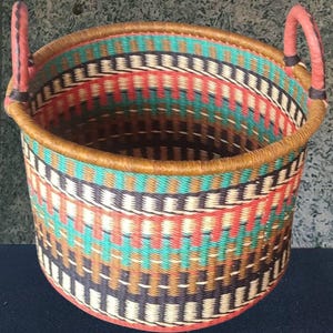 May include: A round woven basket with a colorful geometric pattern. The basket features a combination of red, teal, brown, black, and cream-colored woven strands. It has two pink handles and a brown leather trim around the top.