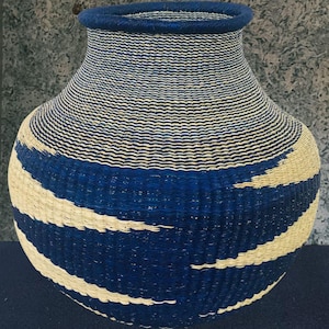 May include: A handwoven basket with a rounded base and a narrow neck. The basket has a pattern of navy blue and cream horizontal stripes. The rim and top of the neck are navy blue.
