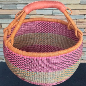 May include: A round woven basket with a pink, orange, and green striped pattern. The basket has a sturdy orange rim and a matching handle with a leather grip. The basket is designed for carrying items.