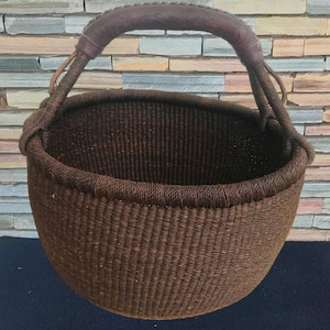 以下が含まれることがあります： 丸みを帯びた形とハンドルが付いた、織り込まれた茶色のバスケット。ハンドルは濃い茶色の革で包まれています。バスケットは、密に織られた繊維でできており、質感のある表面を作り出しています。バスケットはアイテムを運ぶように設計されています。
