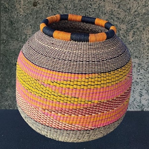 Puede incluir: Una cesta tejida a mano con forma redonda y una abertura ancha. La cesta presenta un diseño vibrante con rayas horizontales en tonos rosa, amarillo, naranja y marrón. El borde está acentuado con bandas naranjas y azul marino.