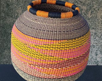 May include: A handwoven basket with a round shape and a wide opening. The basket features a vibrant design with horizontal stripes in shades of pink, yellow, orange, and brown. The rim is accented with orange and navy blue bands.