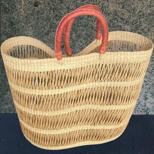 Pode incluir: Cesto grande tecido em bege natural com alça vermelha. O cesto é feito de material tecido e tem um interior espaçoso. As alças vermelhas oferecem um contraste marcante com os tons naturais do cesto.