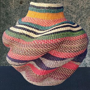 May include: A handwoven basket with a distinctive, twisted form. The basket displays horizontal stripes in various colours, such as red, blue, green, yellow, and brown. The texture of the woven material is evident, enhancing its handcrafted charm.